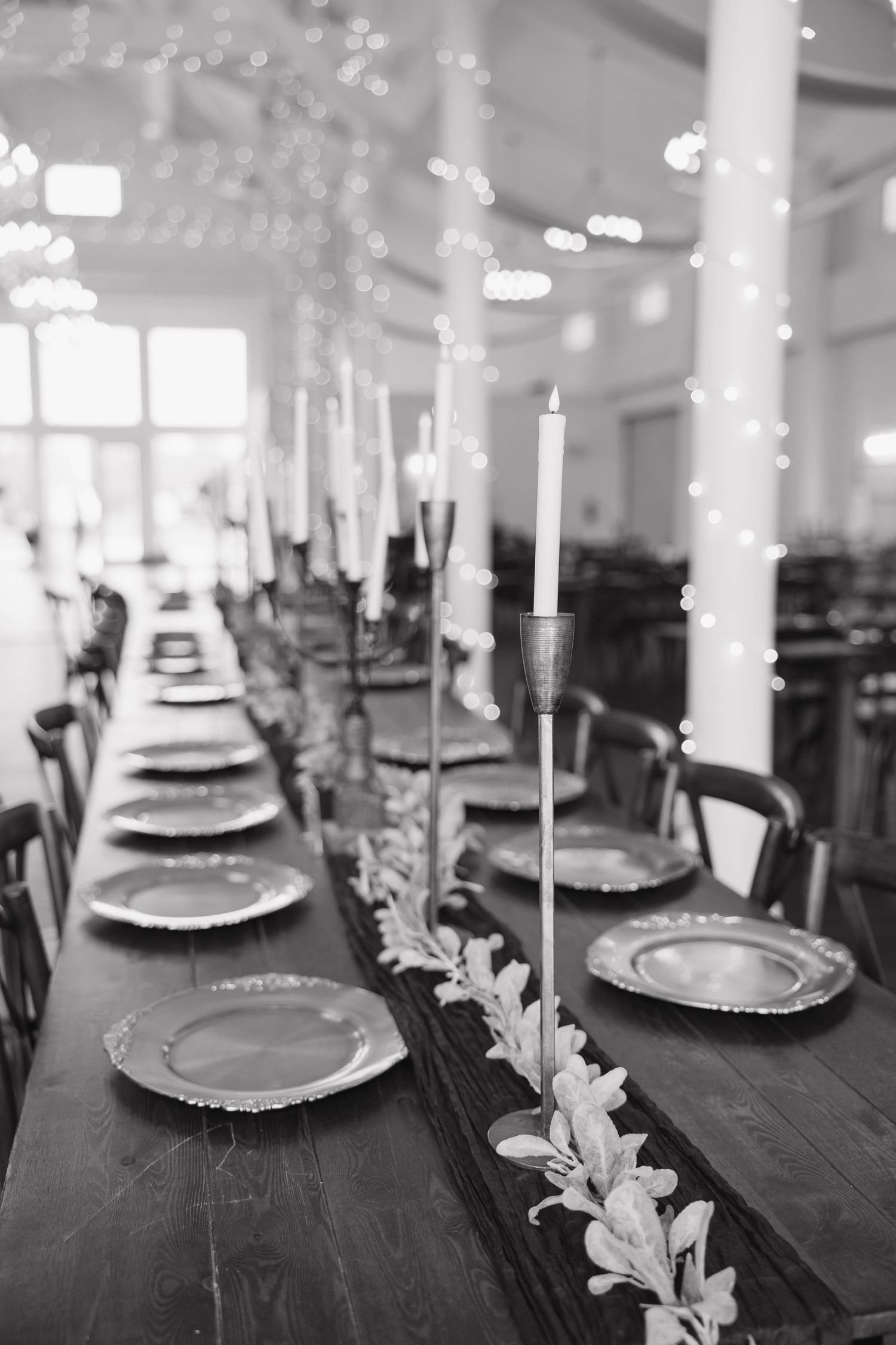 Black and white photo of a long rustic wedding reception table with plates, tall candlesticks, greenery runner and string lights.