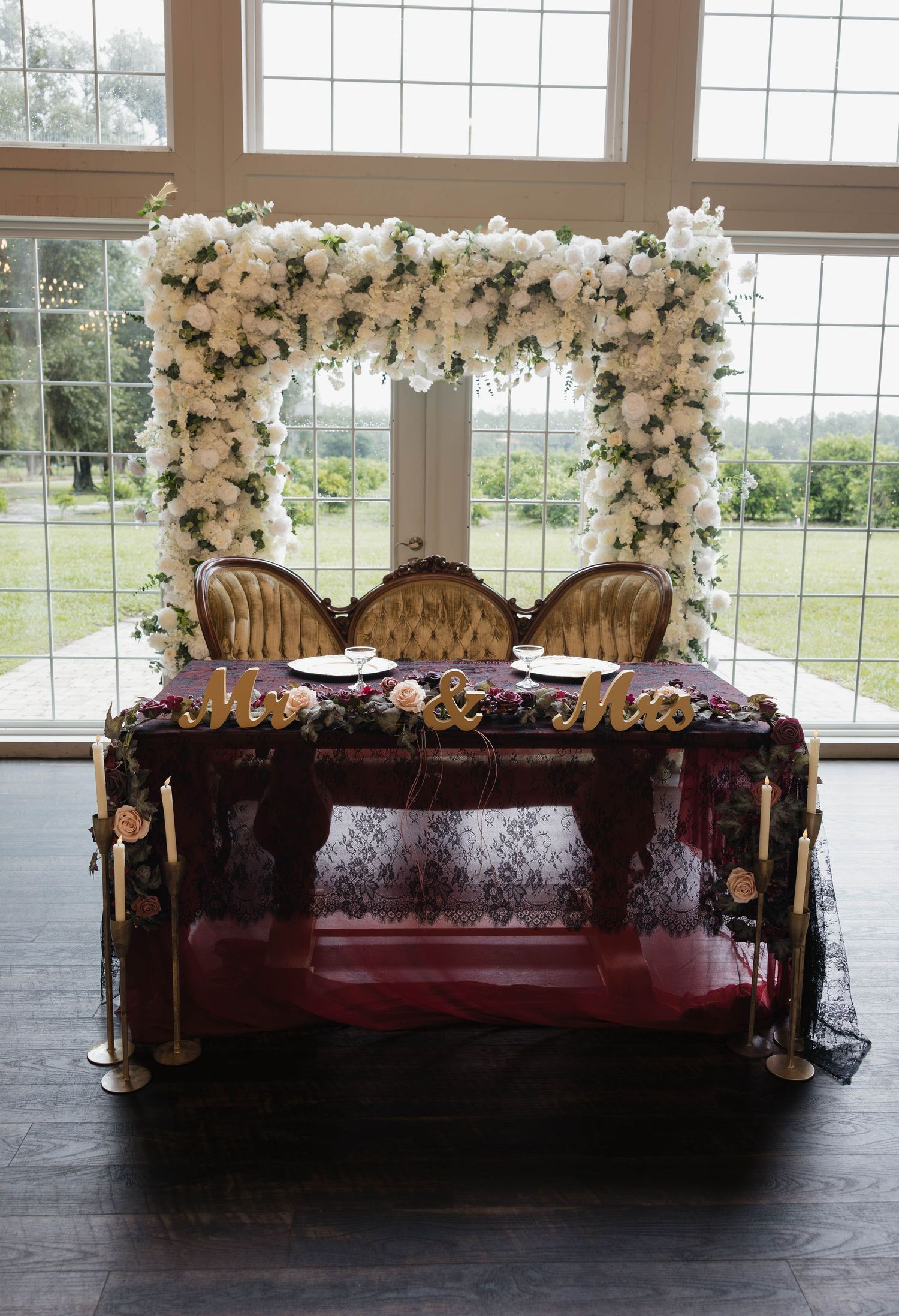 Wedding sweetheart table with a white floral arch, gold Mr and Mrs letters, burgundy linens, and tall candles in a bright venue.