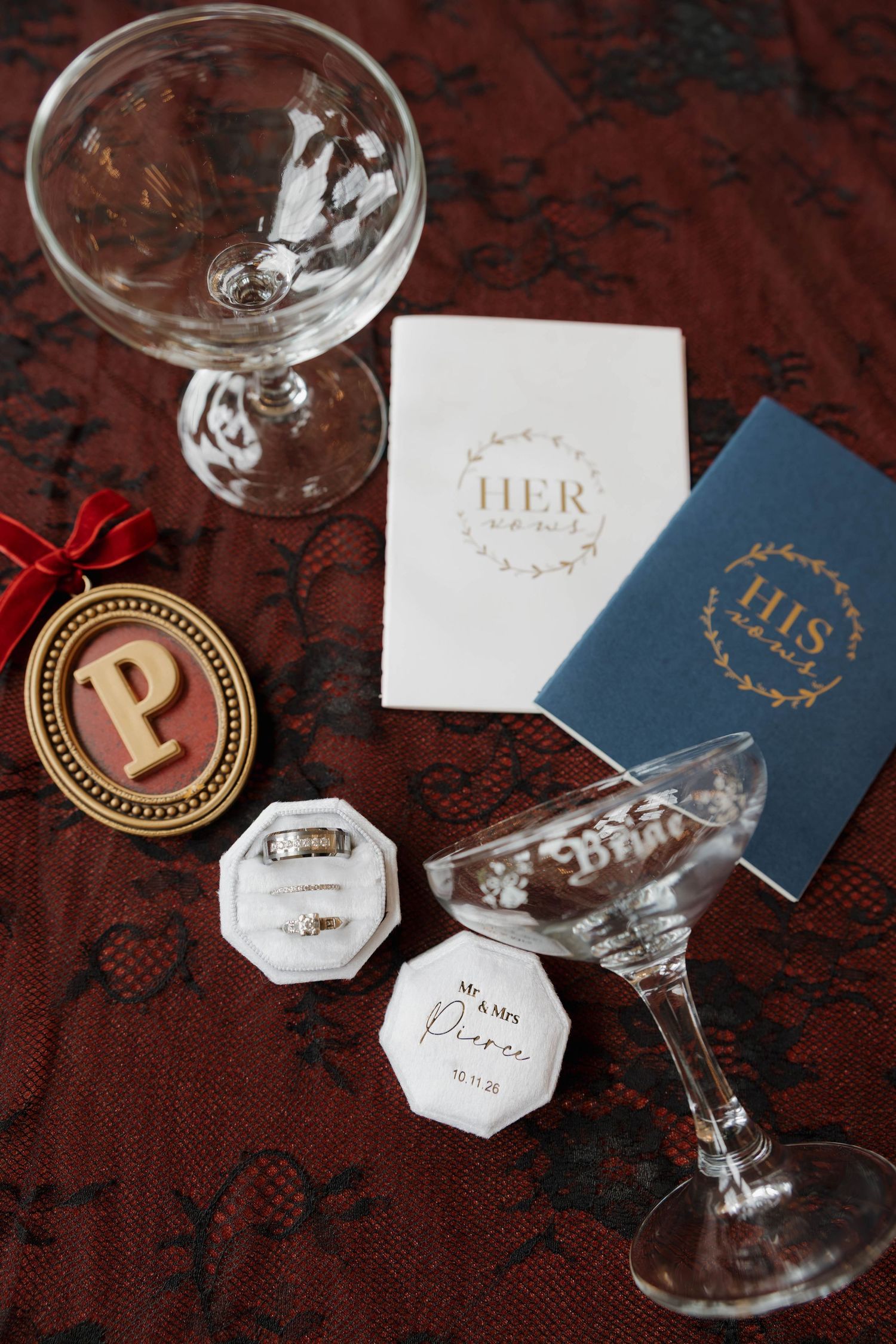 Wedding flat lay with his and hers vow books, coupe glasses, a monogram P ornament, and ring boxes for Mr. & Mrs. Pierce on burgundy fabric.