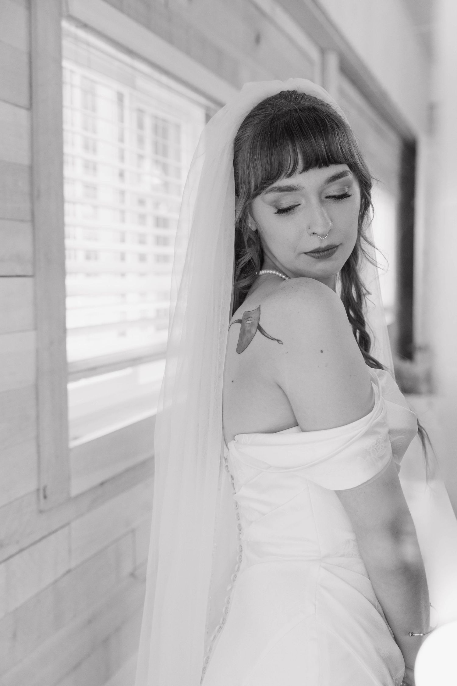 Black and white portrait of a bride with bangs and a long veil, eyes closed, wearing an off-shoulder gown by a sunlit window.