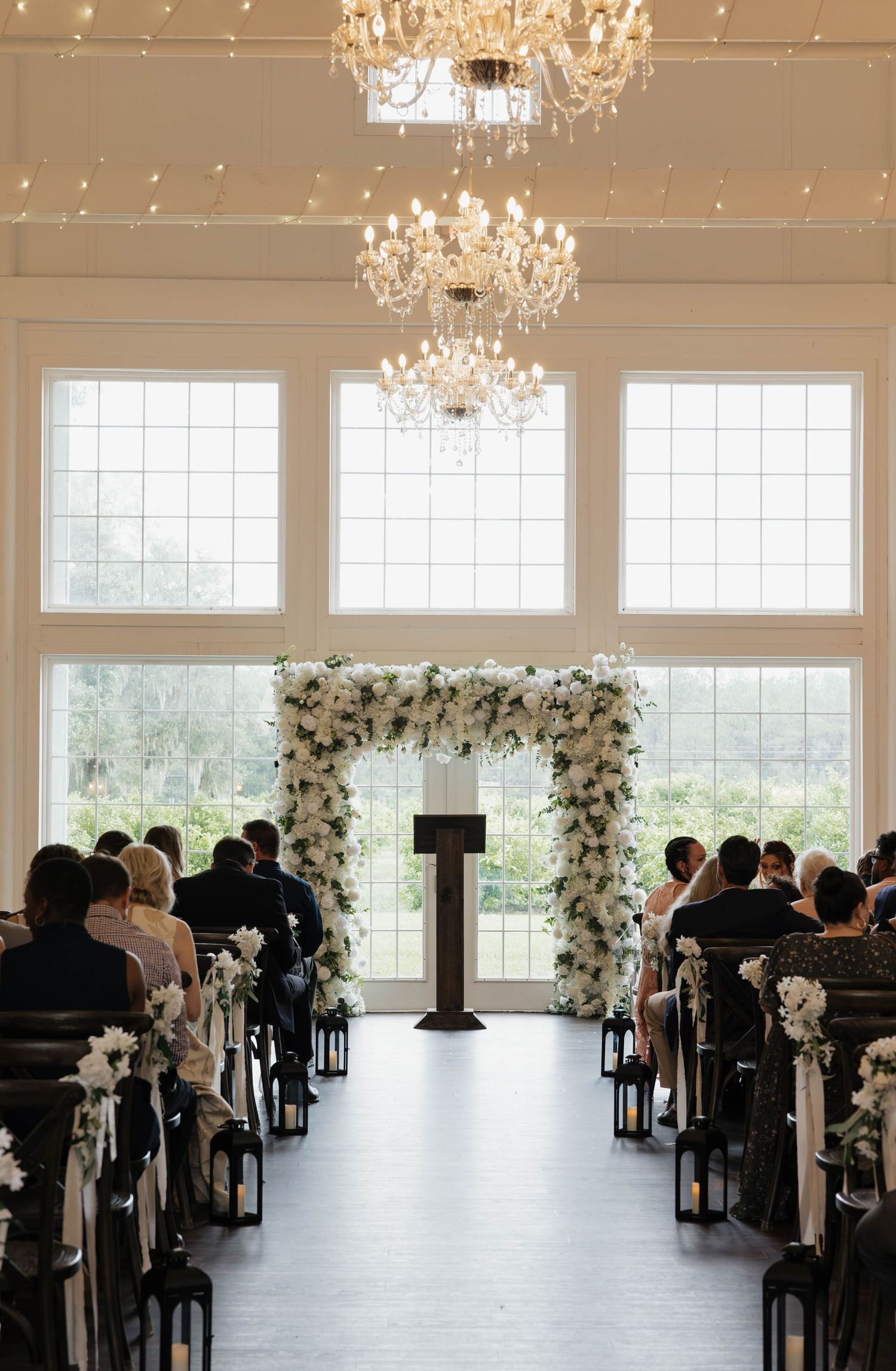 Wedding ceremony space with guests seated facing a white floral arch, wooden podium, and crystal chandeliers beside tall windows.