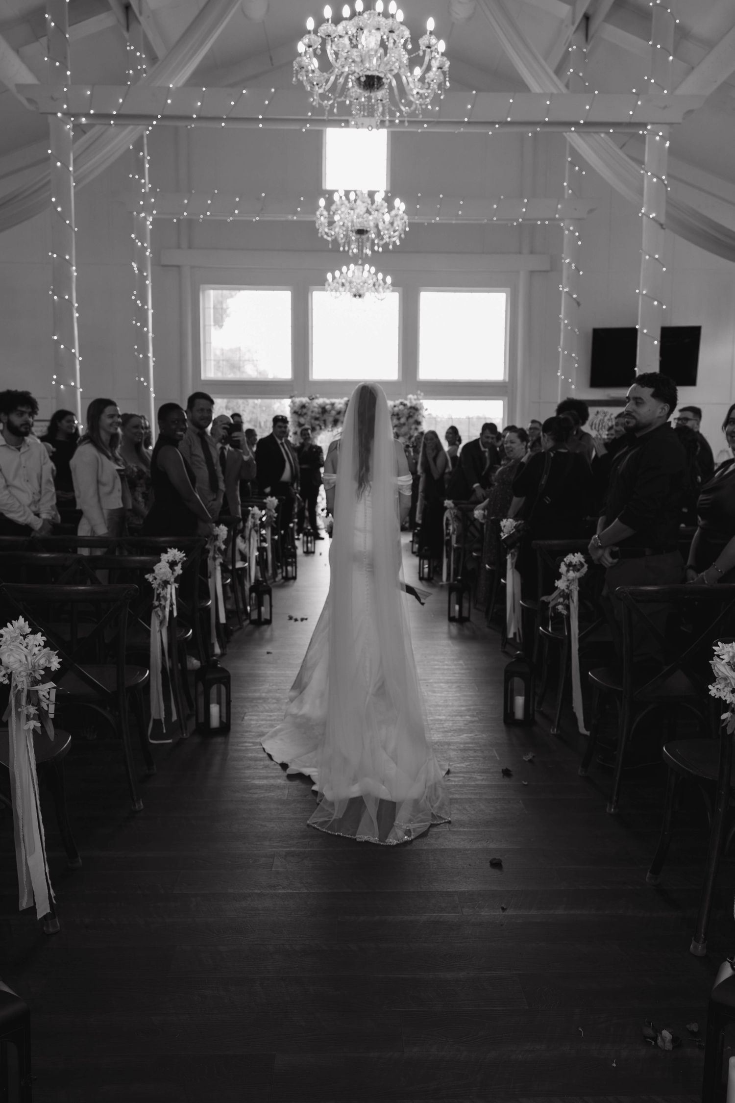 Black and white photo of a bride in a long veil and flowing gown walking down the aisle toward guests under glowing chandeliers.