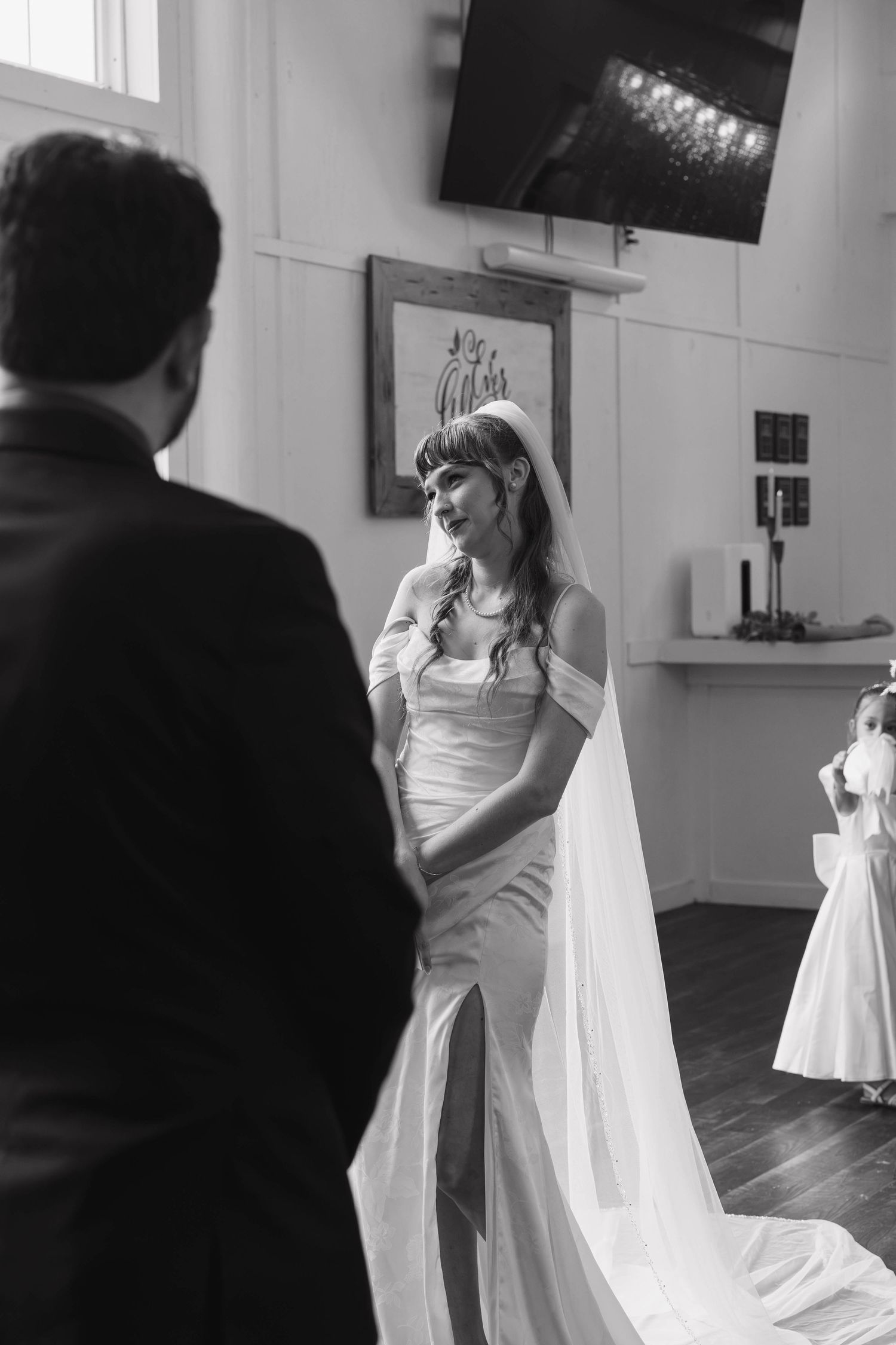 Black and white photo of a tearful bride in a long veil and satin gown facing her groom during the ceremony, flower girl nearby.
