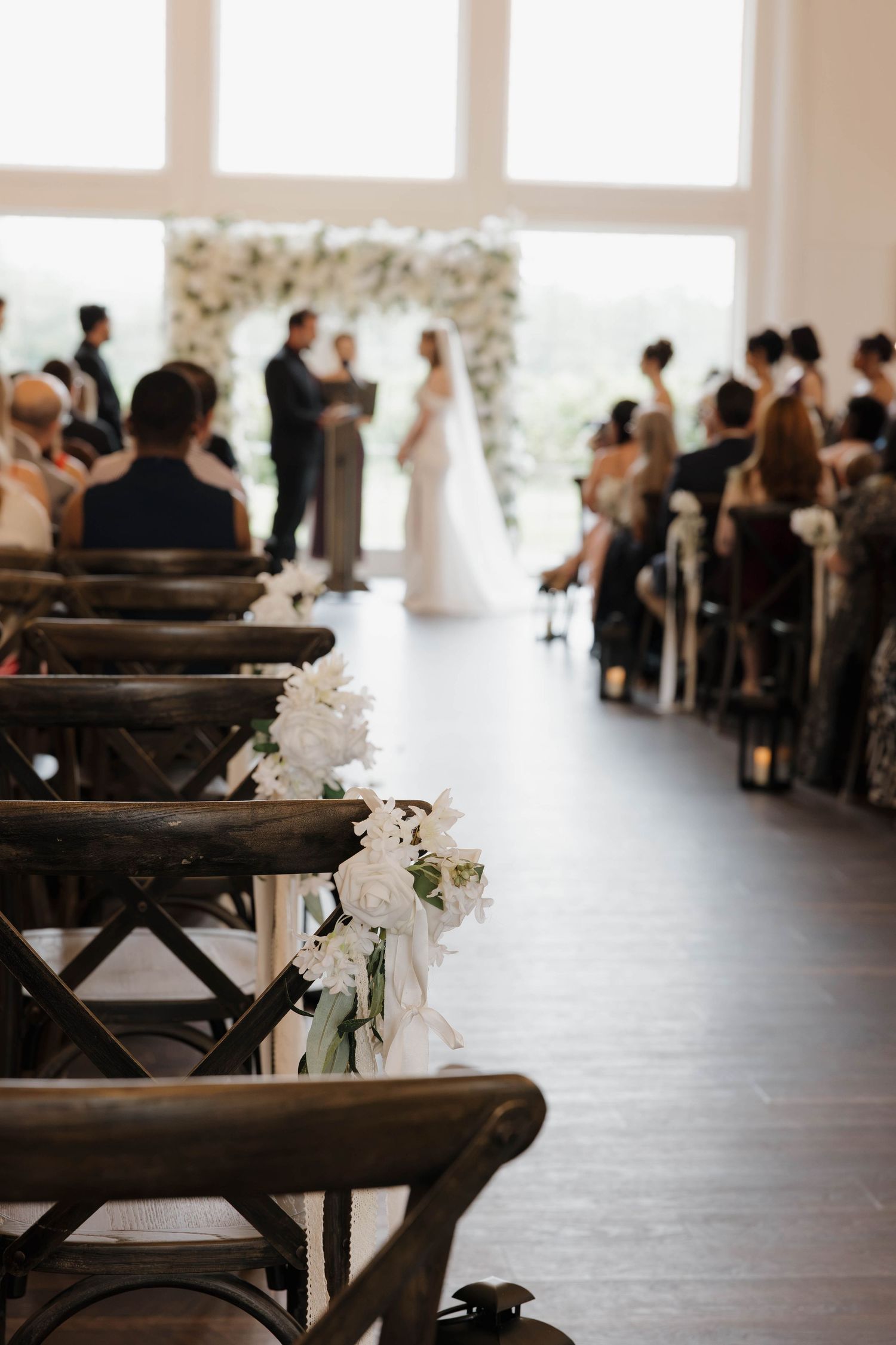 Bride in a white gown stands at a floral arch as wedding guests sit on wooden chairs with white flower arrangements lining the aisle.