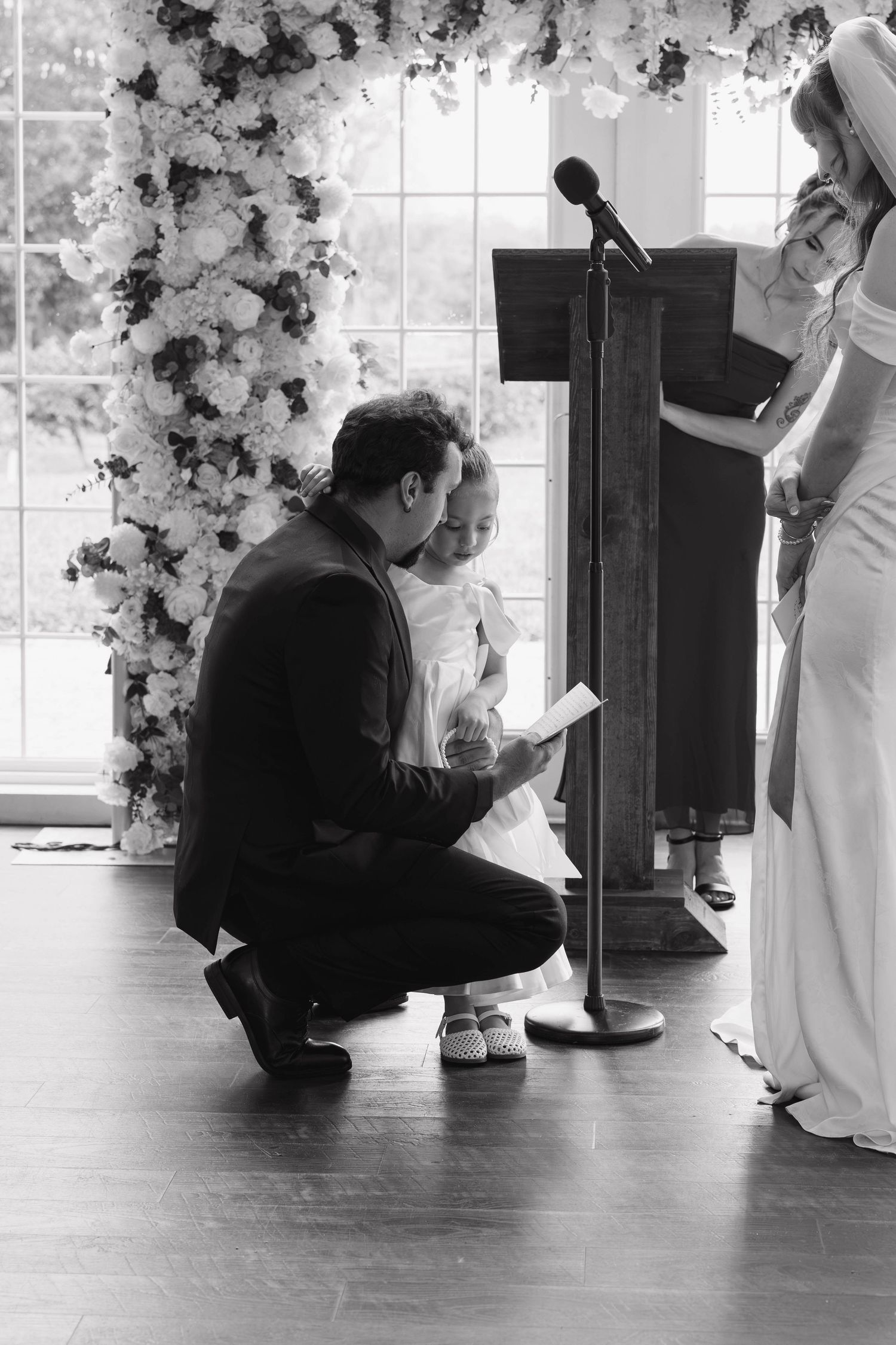 Black and white image of a groom kneeling to read a note to a flower girl beside a floral arch during the wedding ceremony.