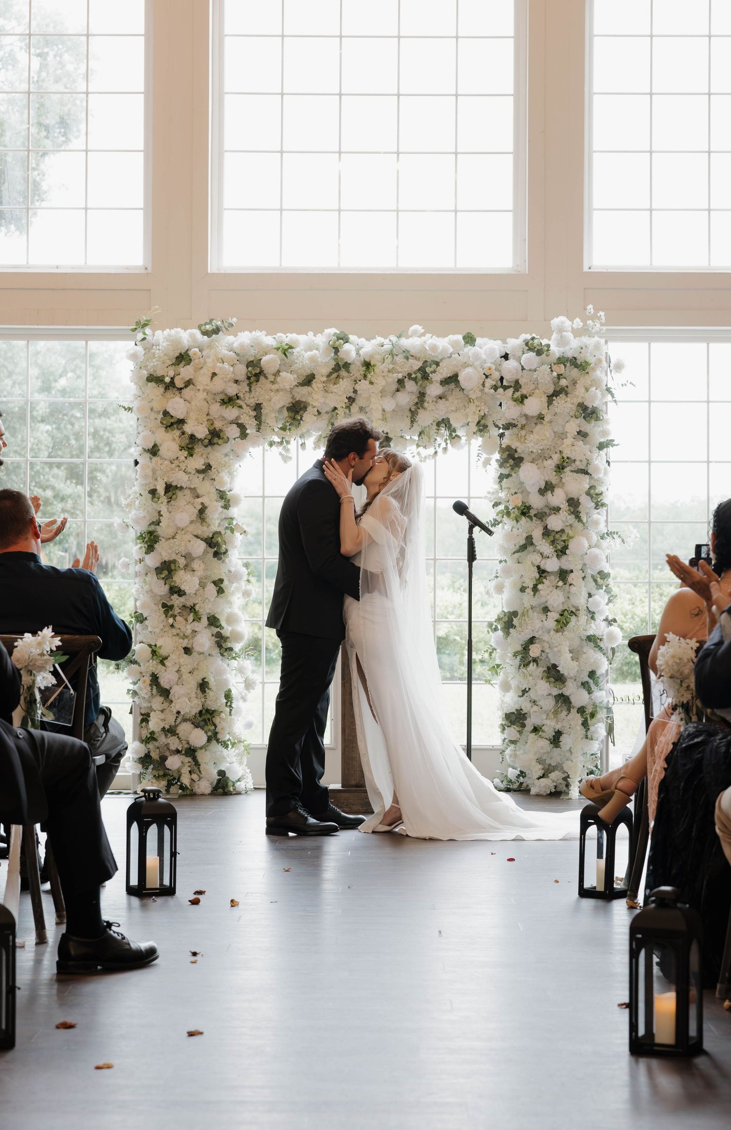 Bride and groom kiss beneath a white floral wedding arch during an indoor ceremony as guests applaud.
