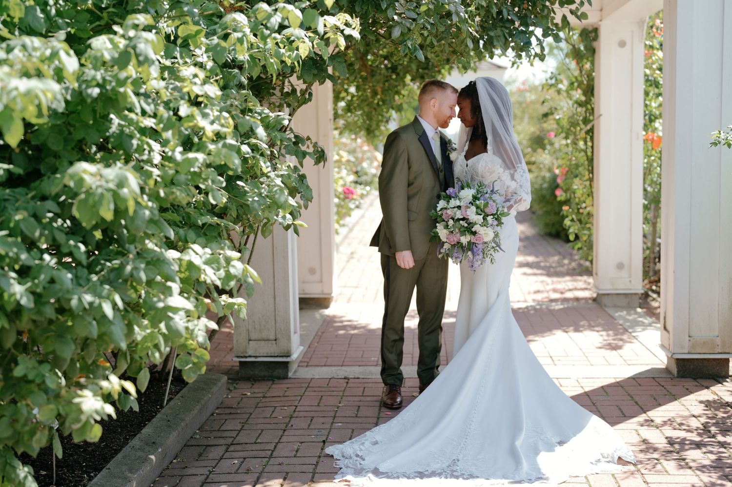 Bride in a flowing white gown and veil holding a bouquet kisses her groom in a green suit on a brick garden path lined with lush greenery.