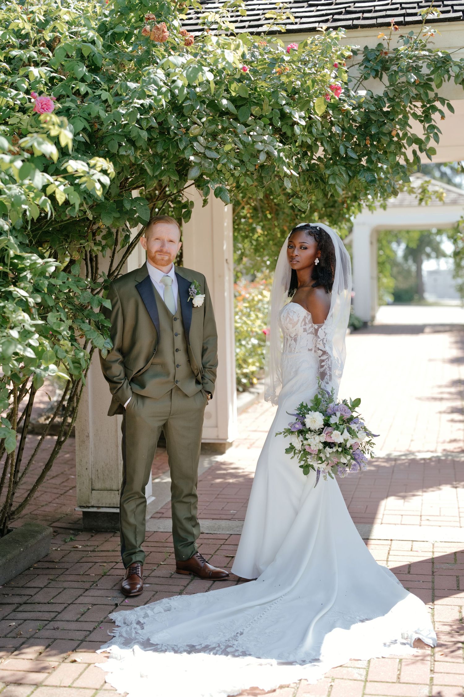 Karen and Yan stand together on a brick path at Dogwood Pavilion, Karen holding her bouquet.