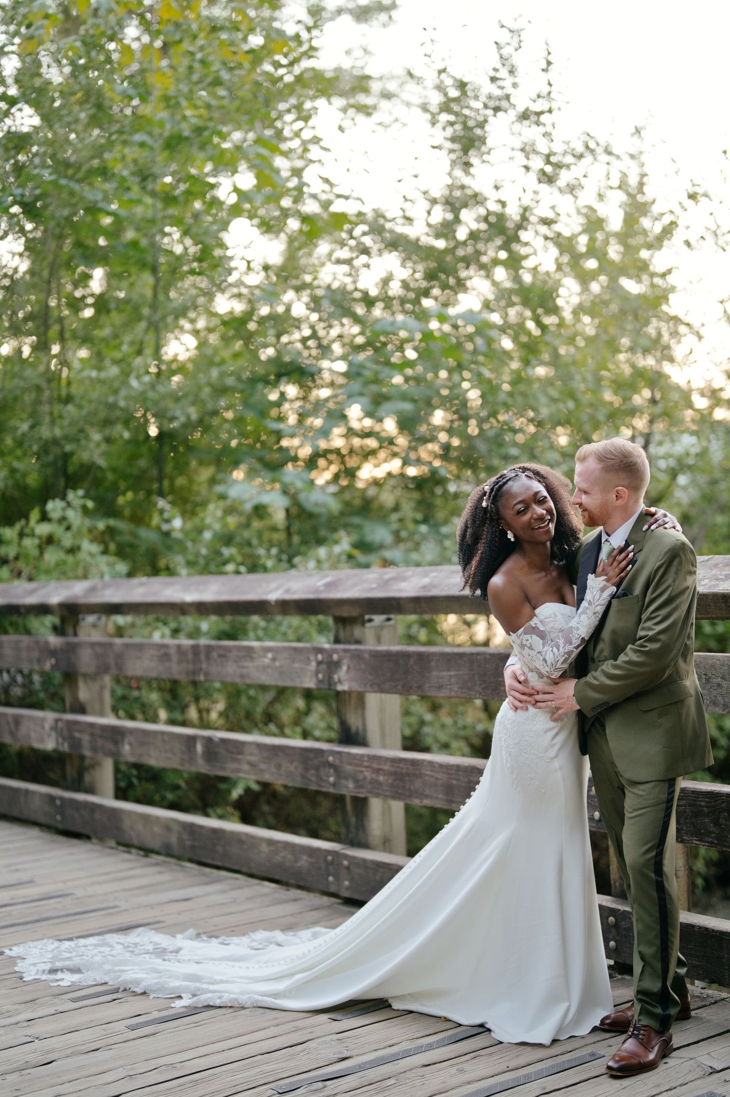 Bride in a lace wedding gown embraces her groom in an olive green suit on a wooden bridge surrounded by lush green trees.