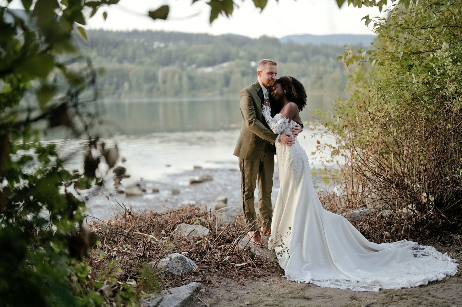 Bride in a flowing white gown embraces her groom in an olive suit on a rocky lakeside shore surrounded by evergreen trees at a wedding.