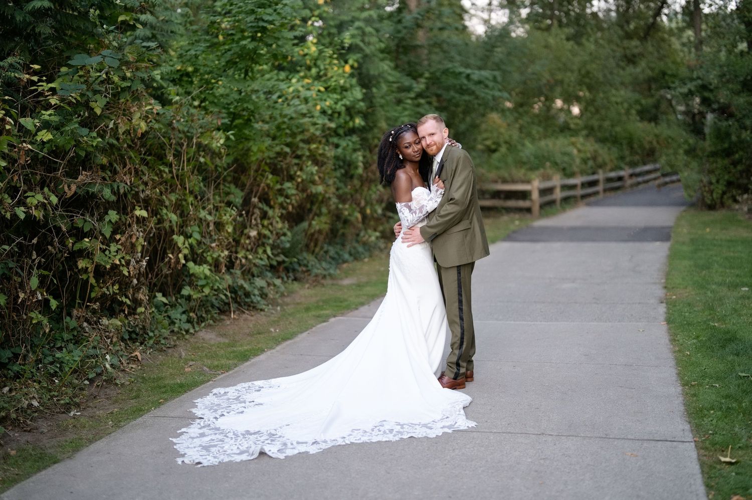 Bride in a long white wedding gown embraces her groom in an olive suit on a paved path surrounded by lush green trees.