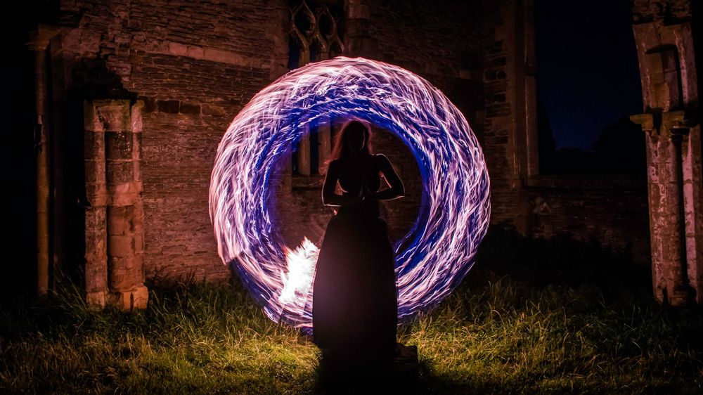 Person performing fire spinning at night creating glowing blue circular light trails near ancient stone ruins.