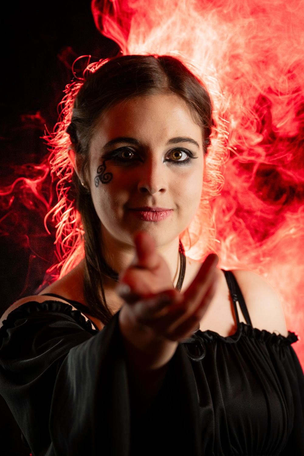 Woman in black dress with dramatic makeup pointing finger, surrounded by red fire and smoke on dark background.