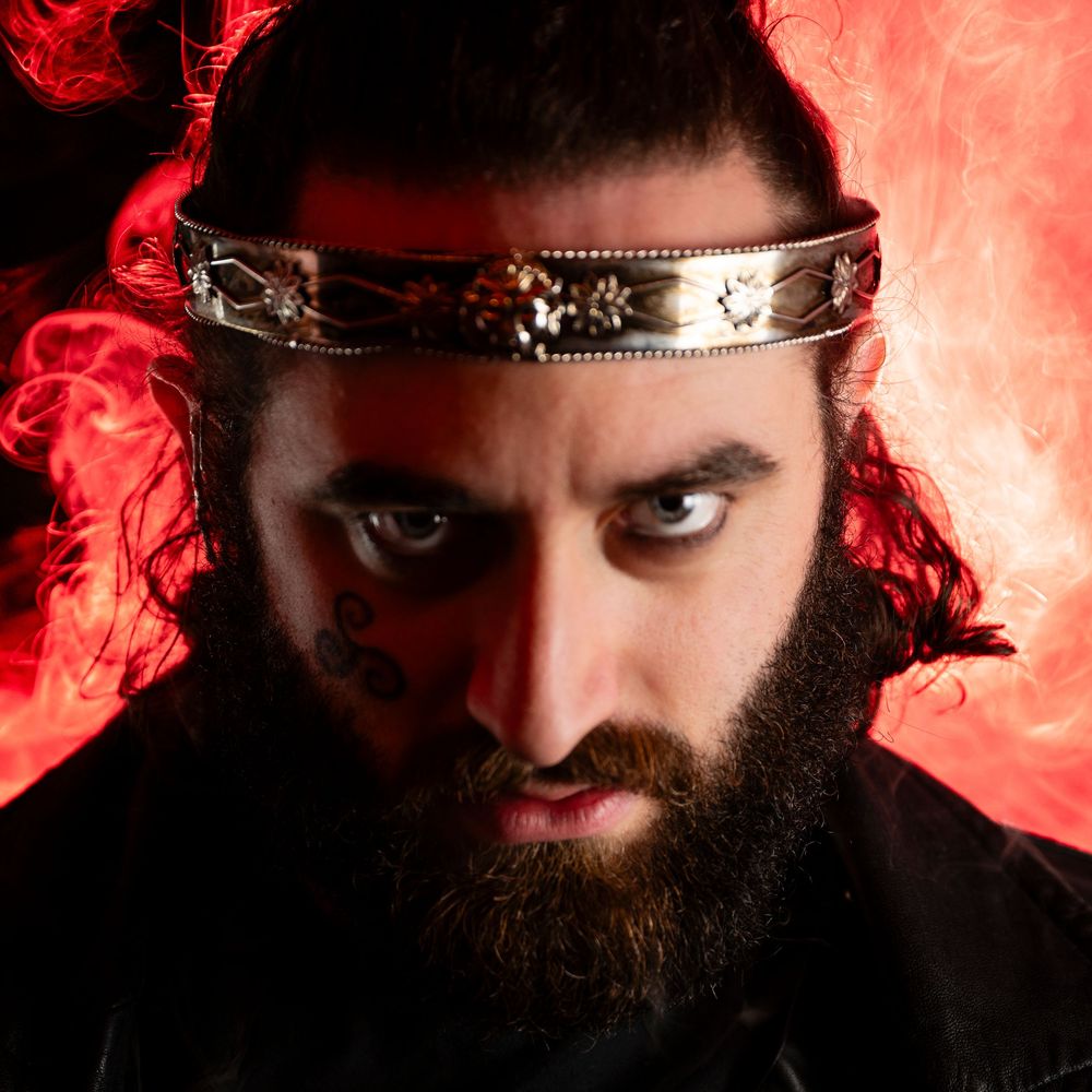 Intense bearded man wearing a decorative headband with face tattoos against a dramatic red smoky background.