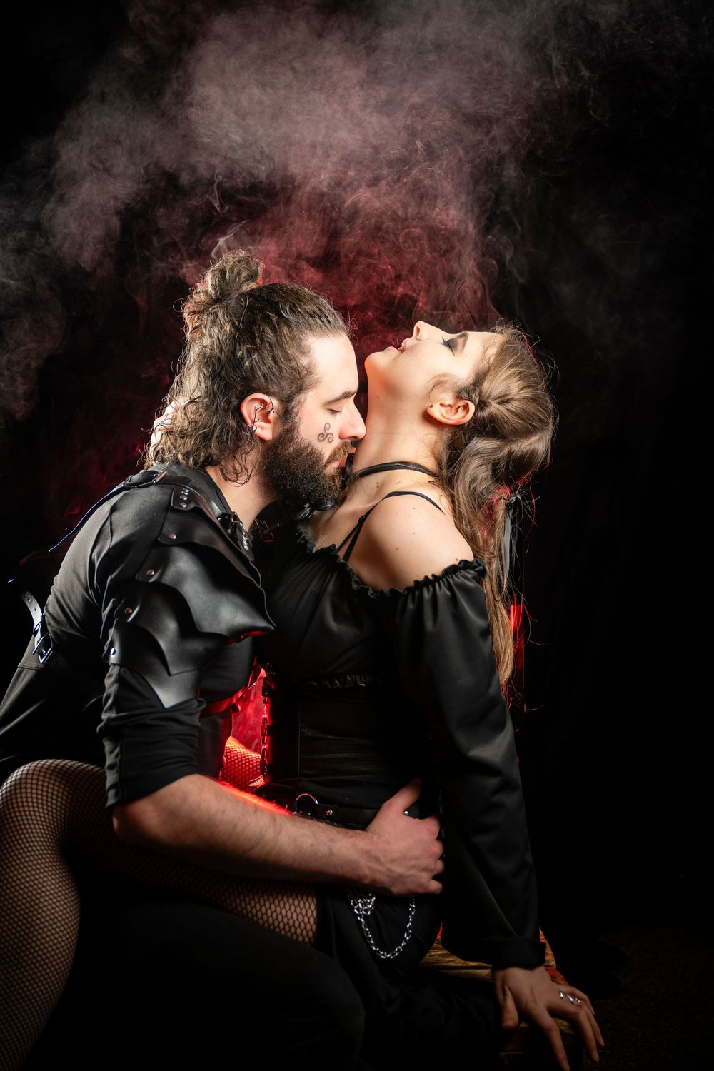 A bearded man in black leather embraces a woman in a dark outfit as they share a passionate kiss against a smoky backdrop.