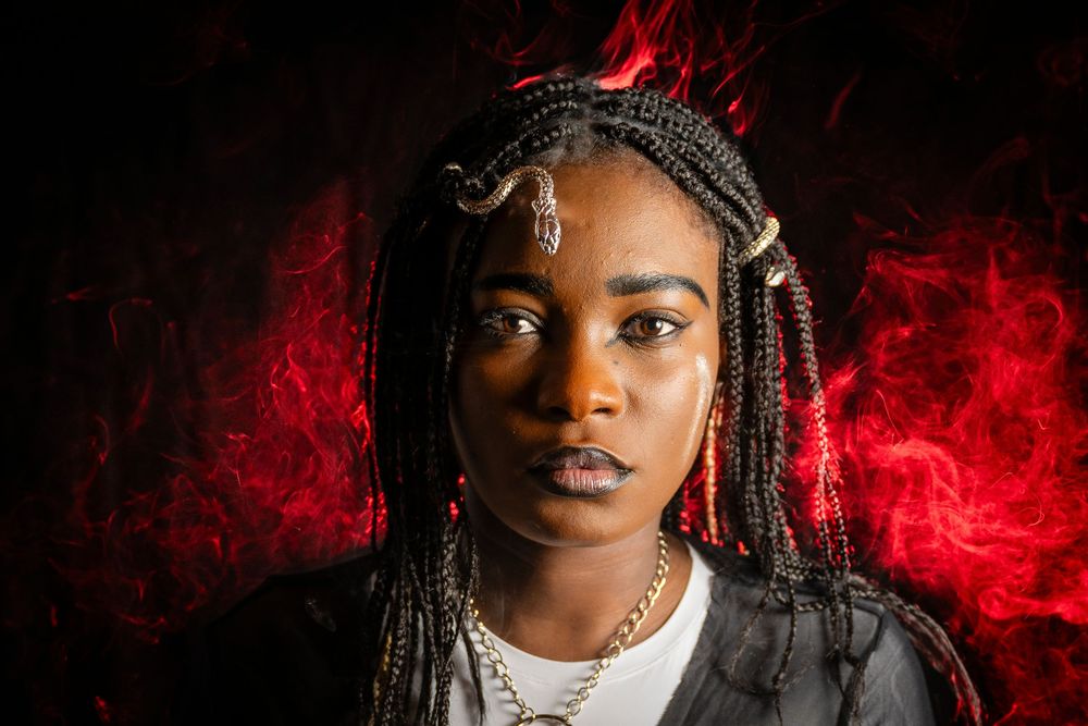 Young woman with braided hair and gold accessories poses against dramatic red smoke background.
