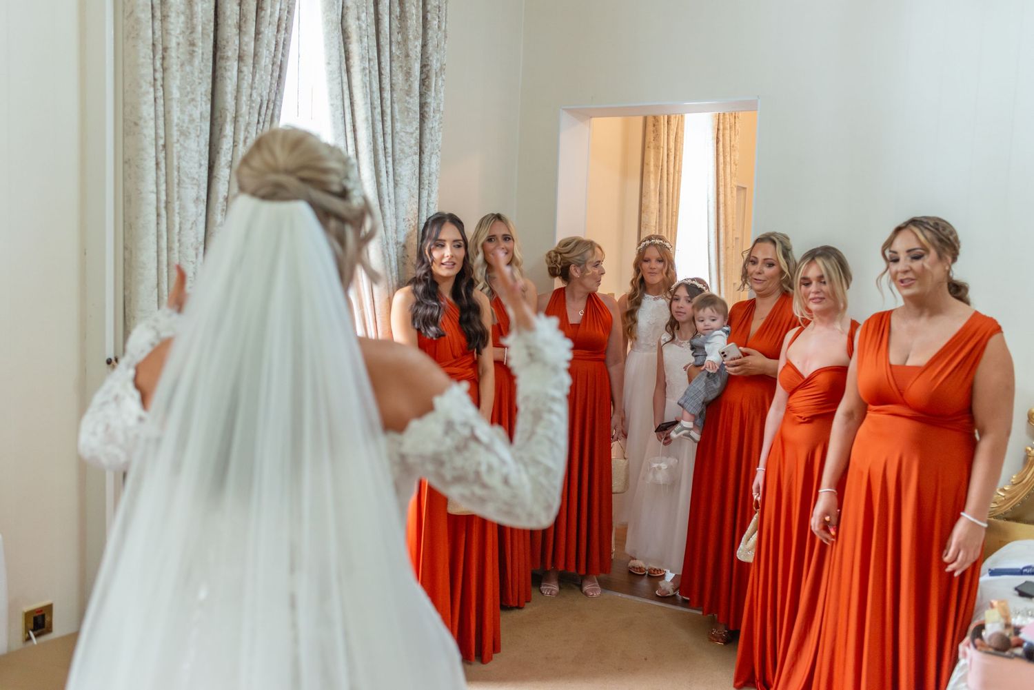 Bride in white gown and veil reveals herself to bridesmaids in matching orange dresses during wedding preparations at Rainhill Hall Hotel
