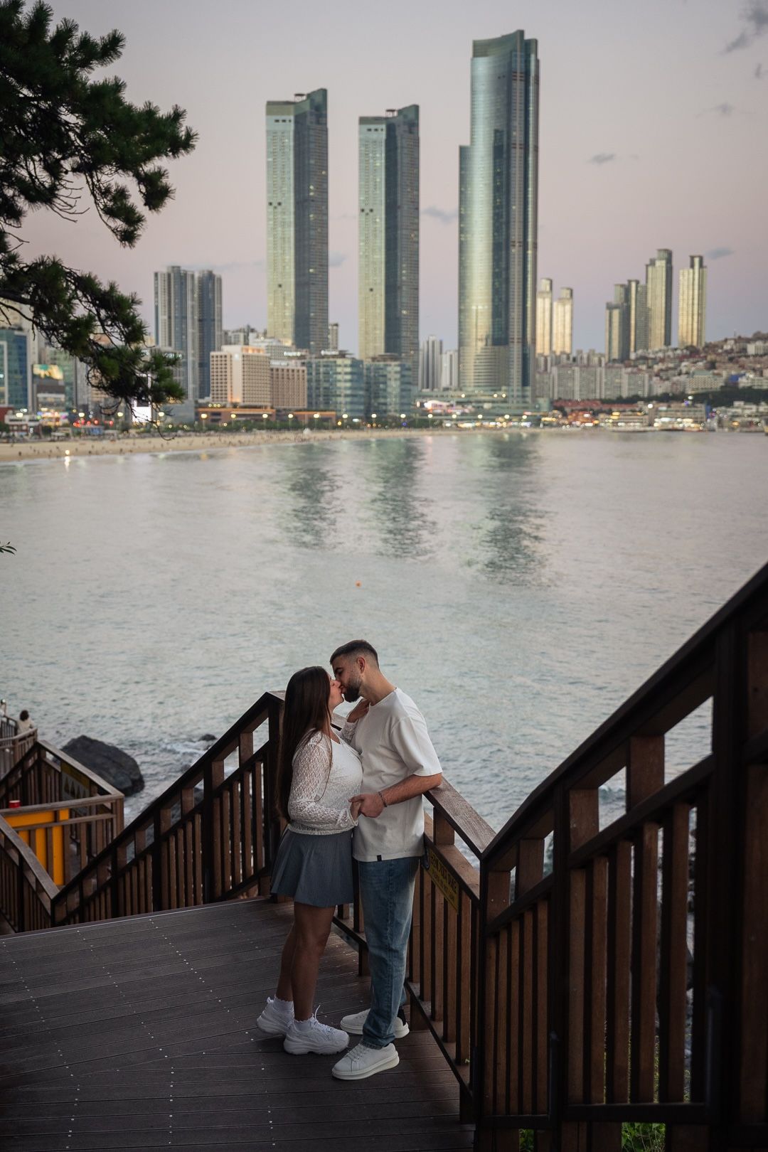 Couple sharing a romantic moment on a wooden deck overlooking Busan city skyline and waterfront at dusk.