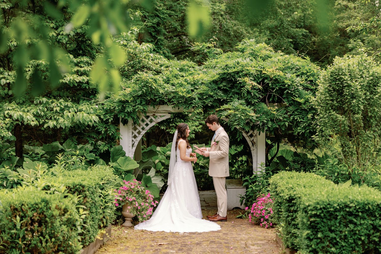 A bride and groom exchange vows under a white garden arbor surrounded by lush green foliage and pink flowers.