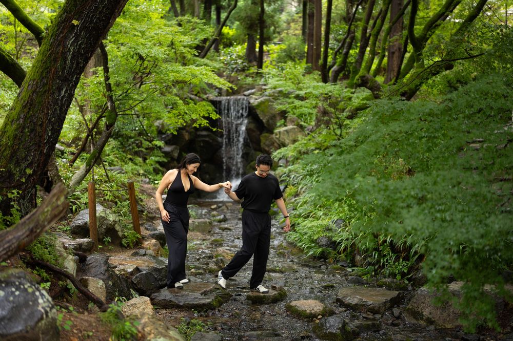 Two people in black clothing explore a lush green forest path beside a serene waterfall on stepping stones.