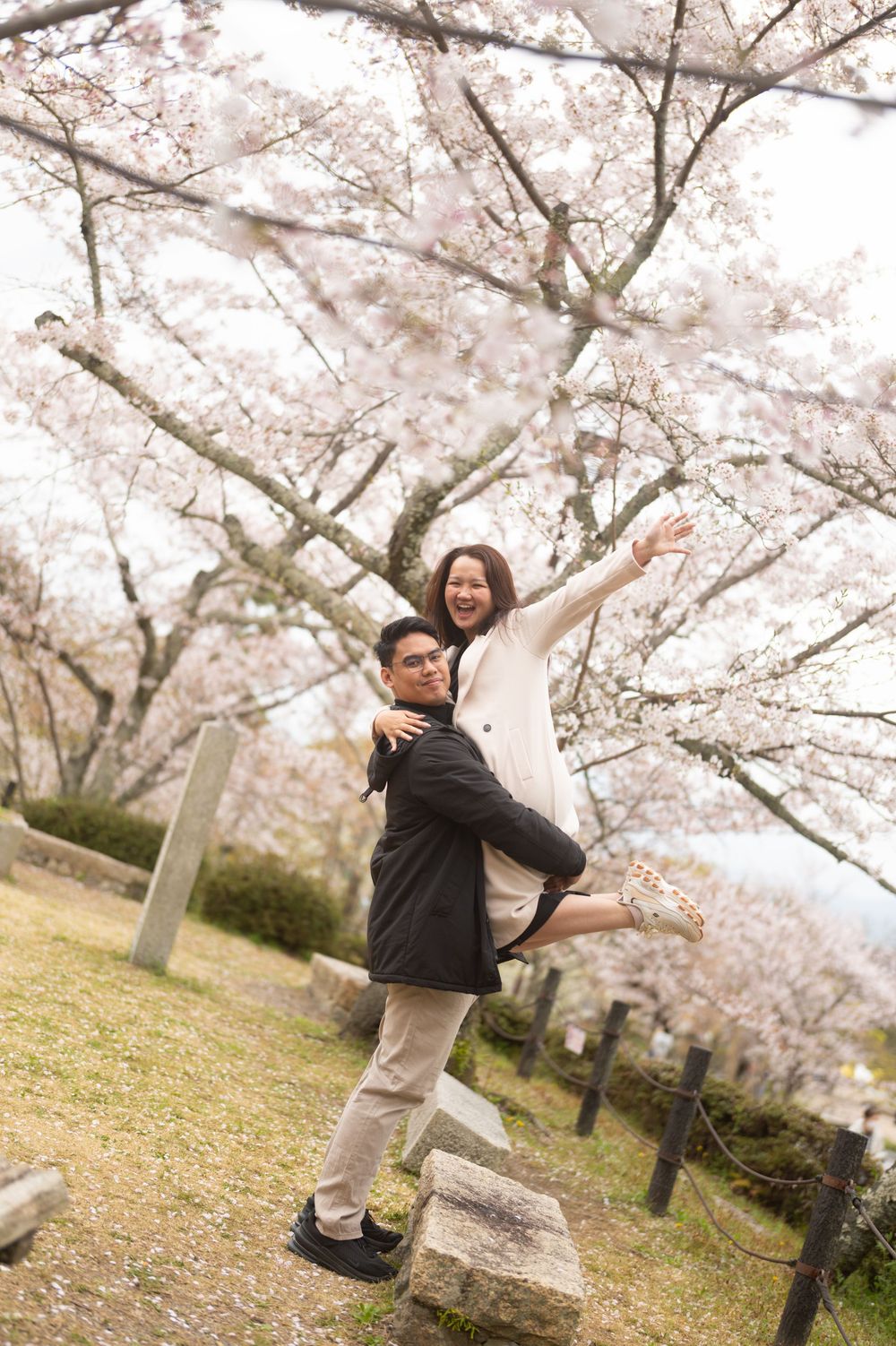 Couple posing joyfully under blooming cherry blossom trees in spring, woman on mans back with arms outstretched.