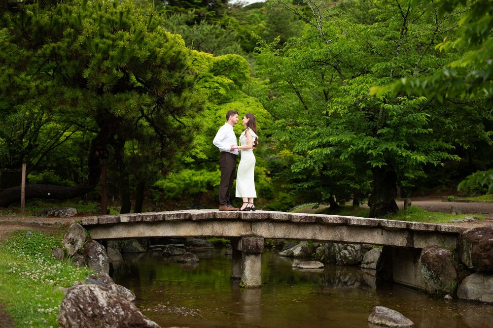 Couple sharing a romantic moment on a stone bridge in a lush Japanese garden with green trees.