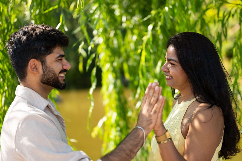 A young couple smiling and greeting each other with a namaste gesture outdoors near lush green willow trees.