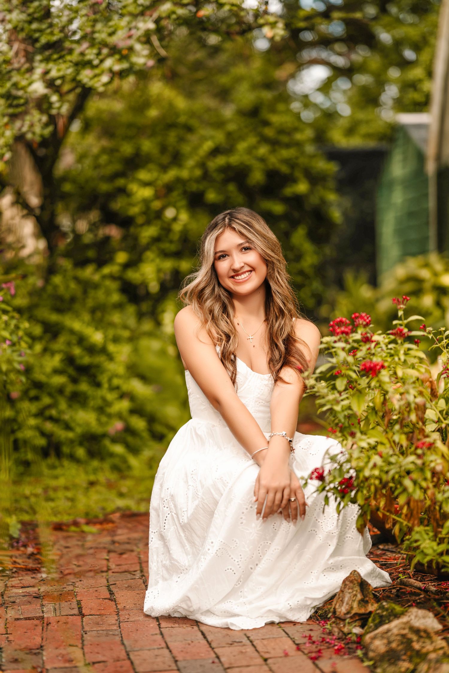 Young woman in white dress sitting on brick path surrounded by lush green garden and red flowers, smiling warmly.
