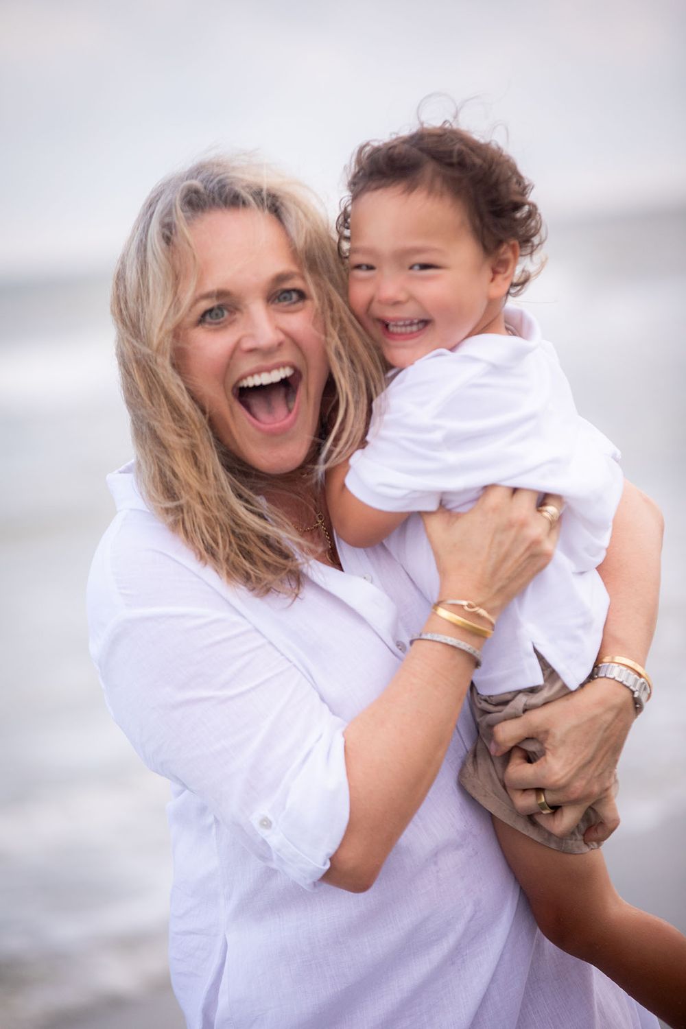A joyful woman in white holds a laughing toddler on a beach, both beaming with happiness near ocean waves.