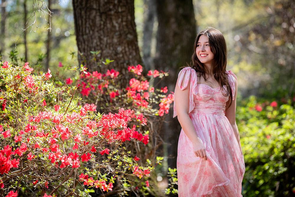 Young woman in pink floral dress smiling among vibrant red azalea bushes in a sunlit garden.