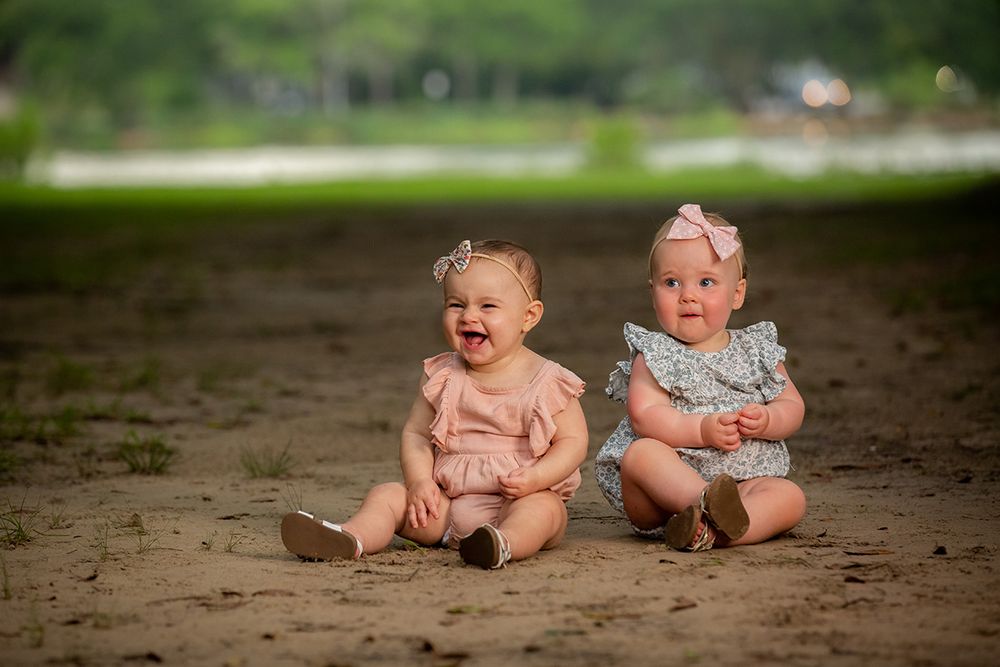 Two adorable baby girls in pink outfits sitting on sandy ground outdoors, smiling and laughing together.