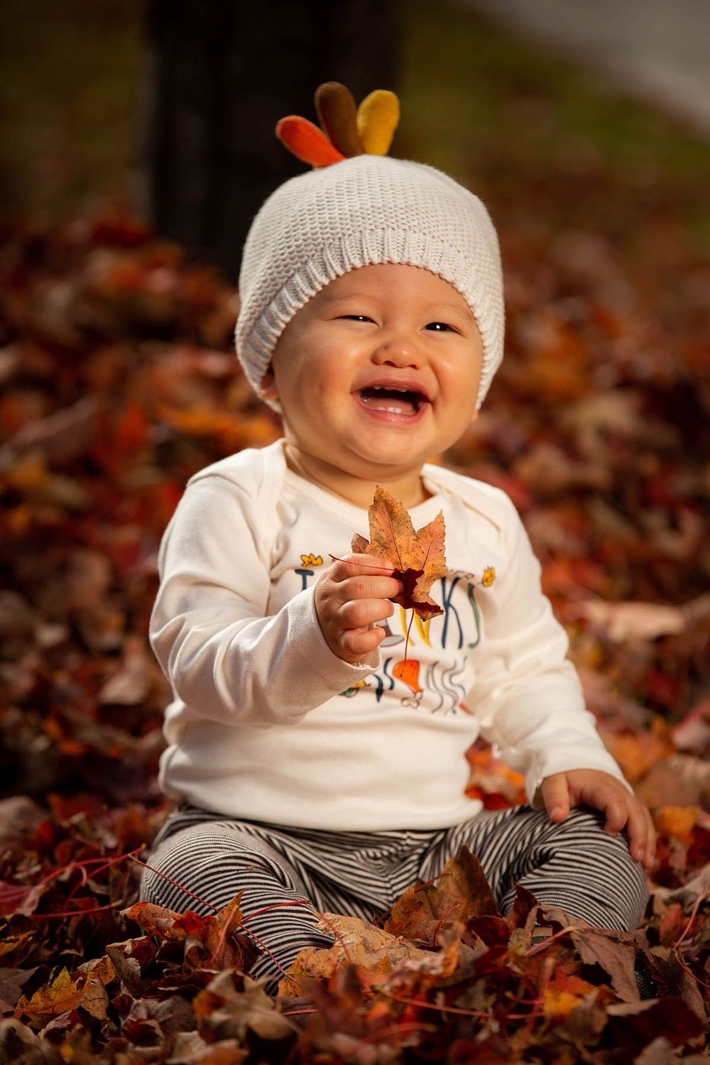 Happy baby in turkey hat holds autumn leaf while sitting in colorful fallen leaves outdoors.