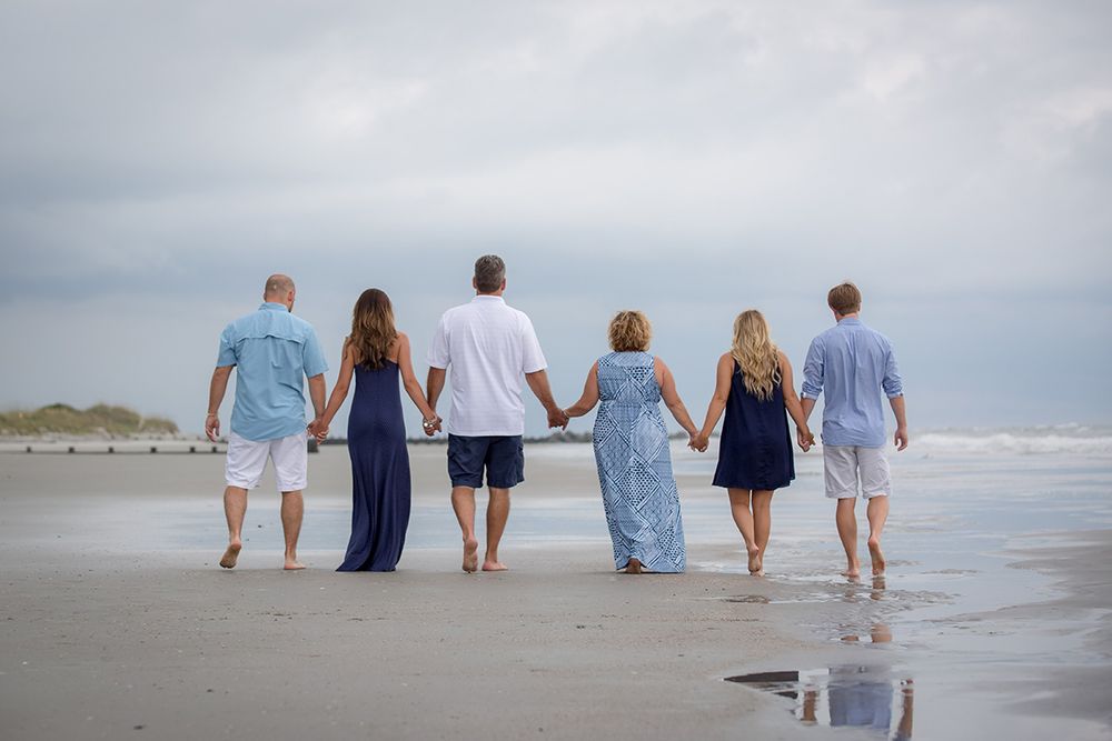 Six people holding hands walk along a sandy beach under cloudy skies, dressed in blue and white outfits.