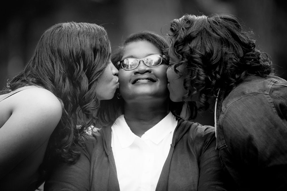 Black and white photo of a smiling woman wearing glasses being kissed on cheeks by two women.