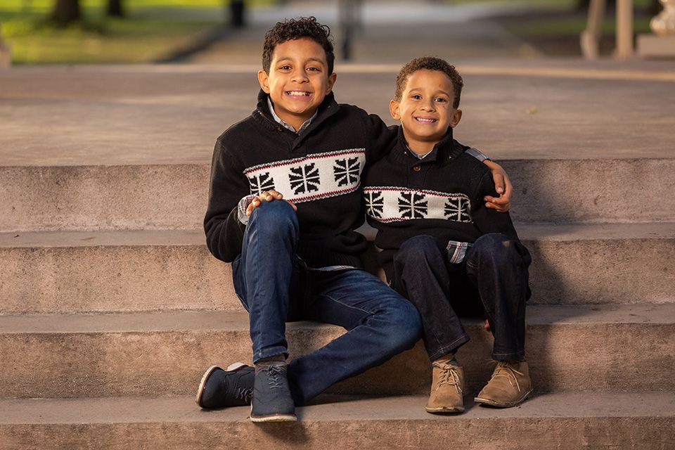 Two smiling boys in matching black Union Jack sweaters and jeans sitting together on stone steps outdoors.
