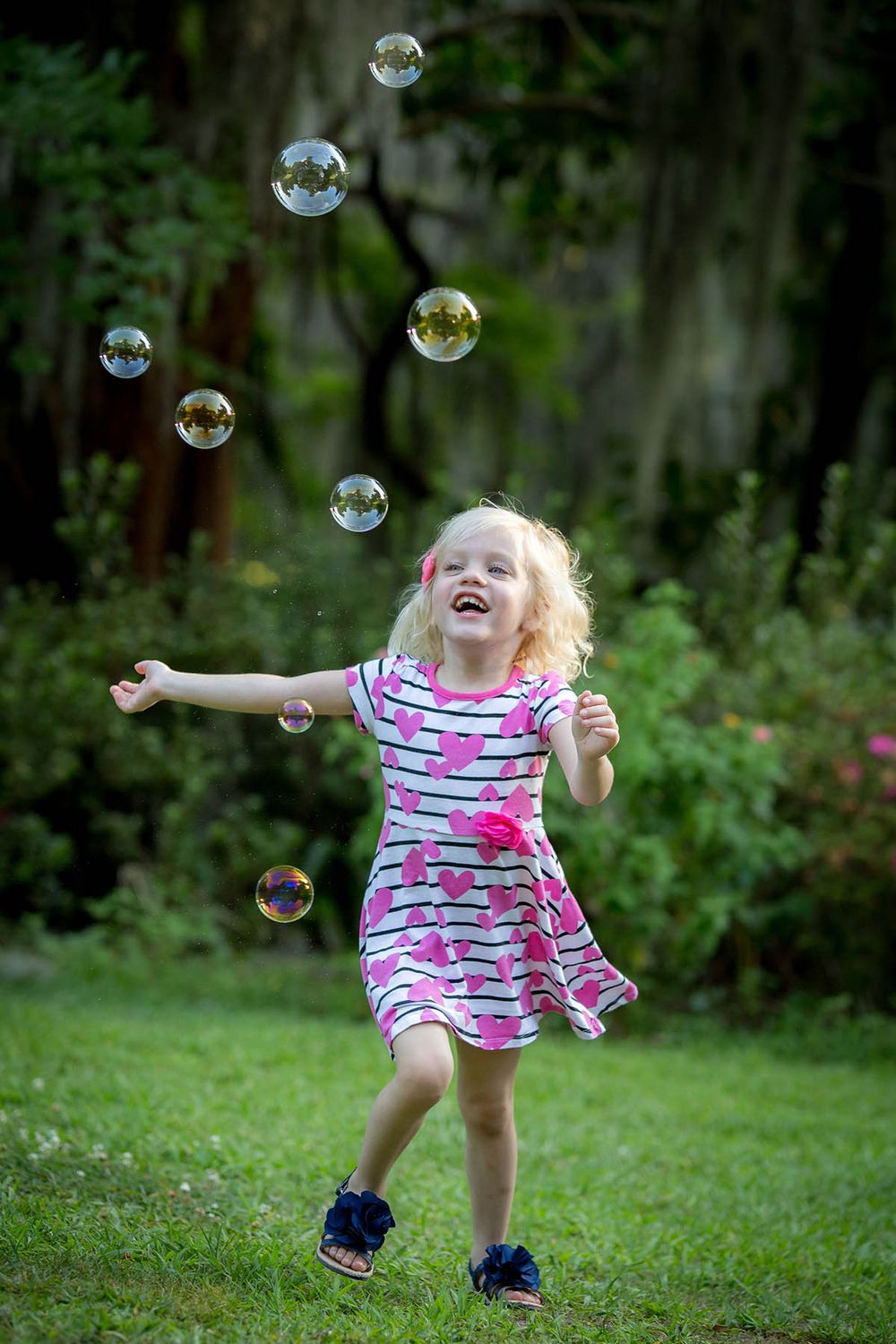 A joyful blonde girl in a pink striped dress laughs while chasing soap bubbles in a lush green backyard.