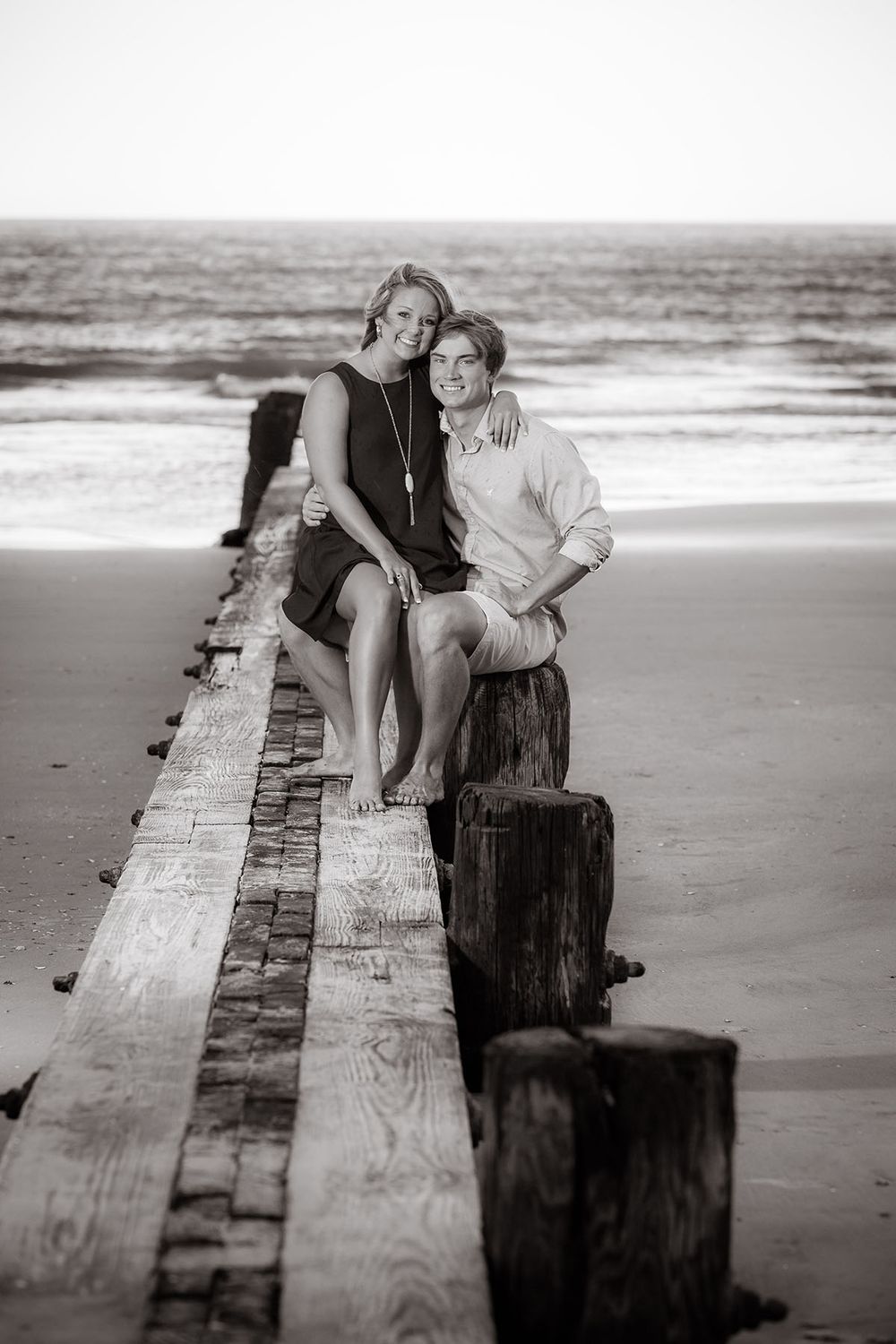 Couple sitting together on weathered wooden pier at beach, black and white romantic seaside portrait photography.