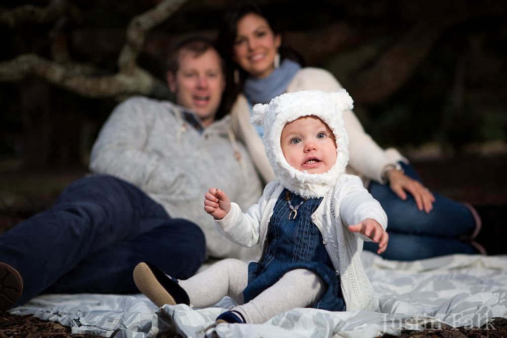 A family with a baby in a white bear hat sits on a blanket outdoors for a professional portrait photo.