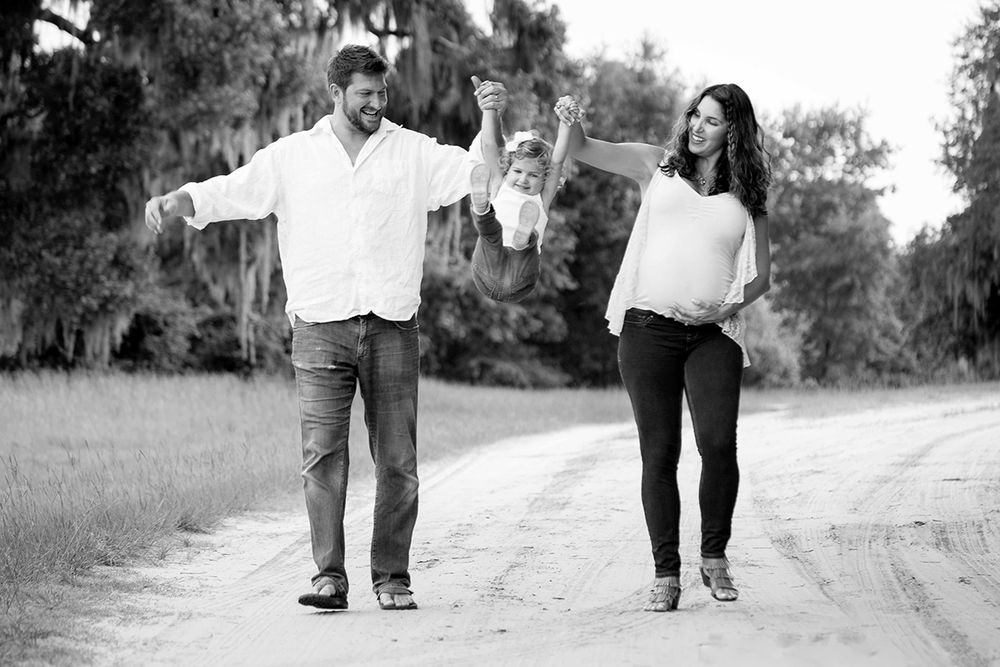 Black and white photo of a couple swinging their toddler on a dirt road, with the woman visibly pregnant.