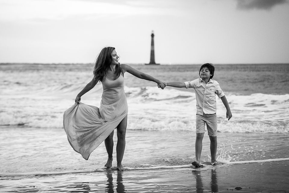 Black and white photo of a woman and child playing on a beach near a lighthouse, holding hands by the ocean waves.