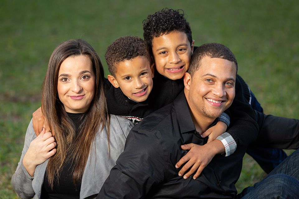 A happy mixed-race family of four smiling outdoors on green grass, with two young boys hugging their parents.