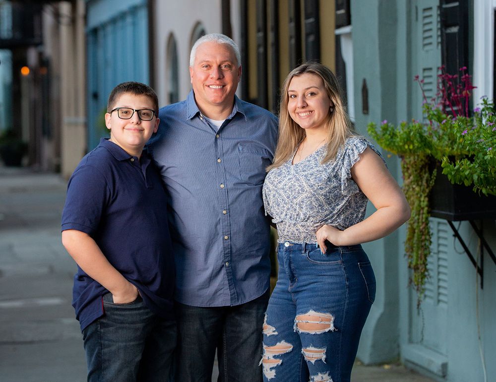 A family of three smiling outdoors on a city sidewalk, wearing casual clothes, posing together for a portrait photo.
