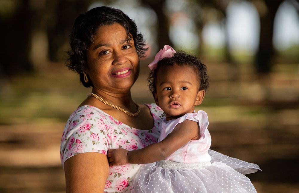 A smiling woman holds a baby girl in a white dress outdoors in a sunlit park setting.