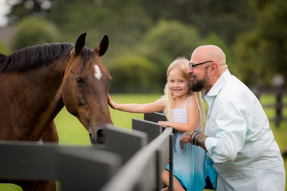A bald man and young blonde girl pet a brown horse over a fence at a lush green farm.