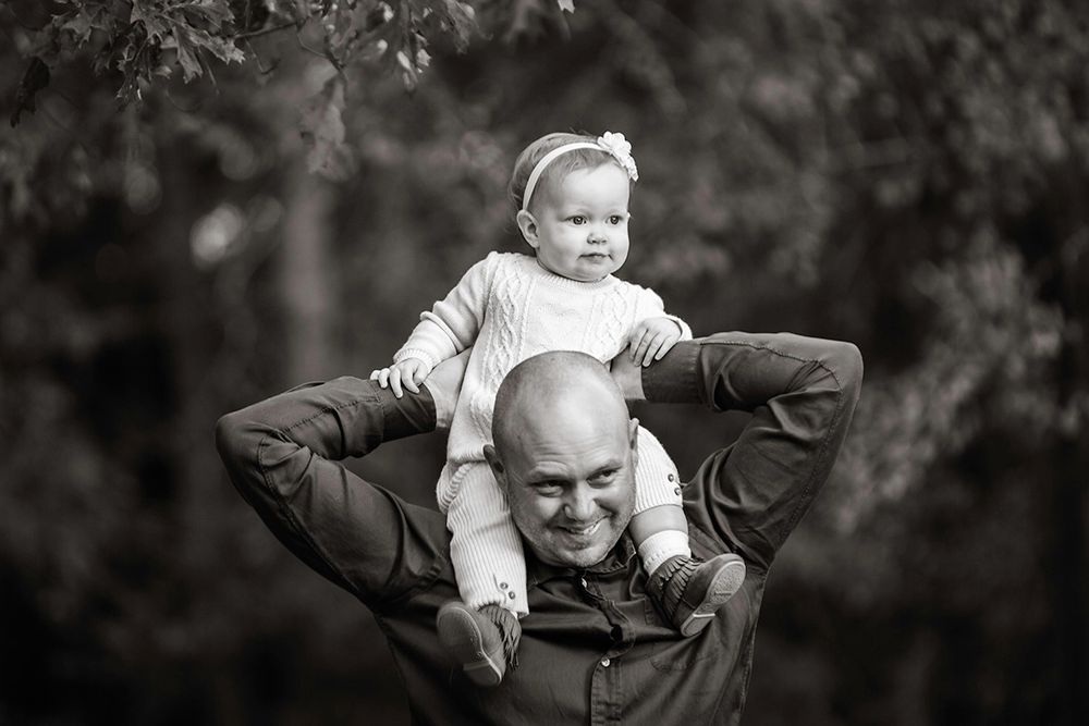 Black and white photo of a bald man smiling with a baby girl sitting on his shoulders outdoors.