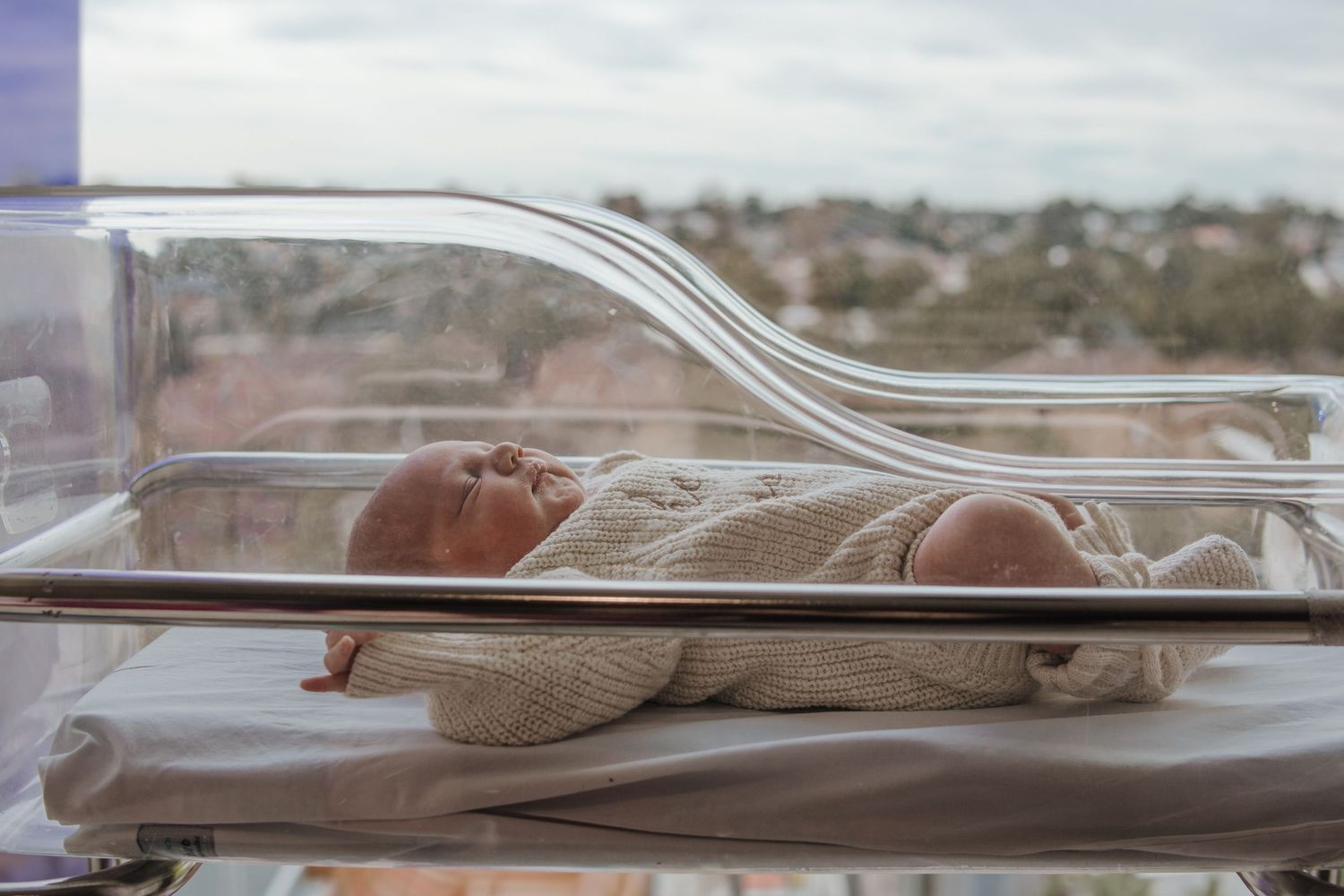 Newborn baby in a cream knit outfit sleeping in a clear hospital bassinet beside a window during hospital fresh 48 session in Blacktown.