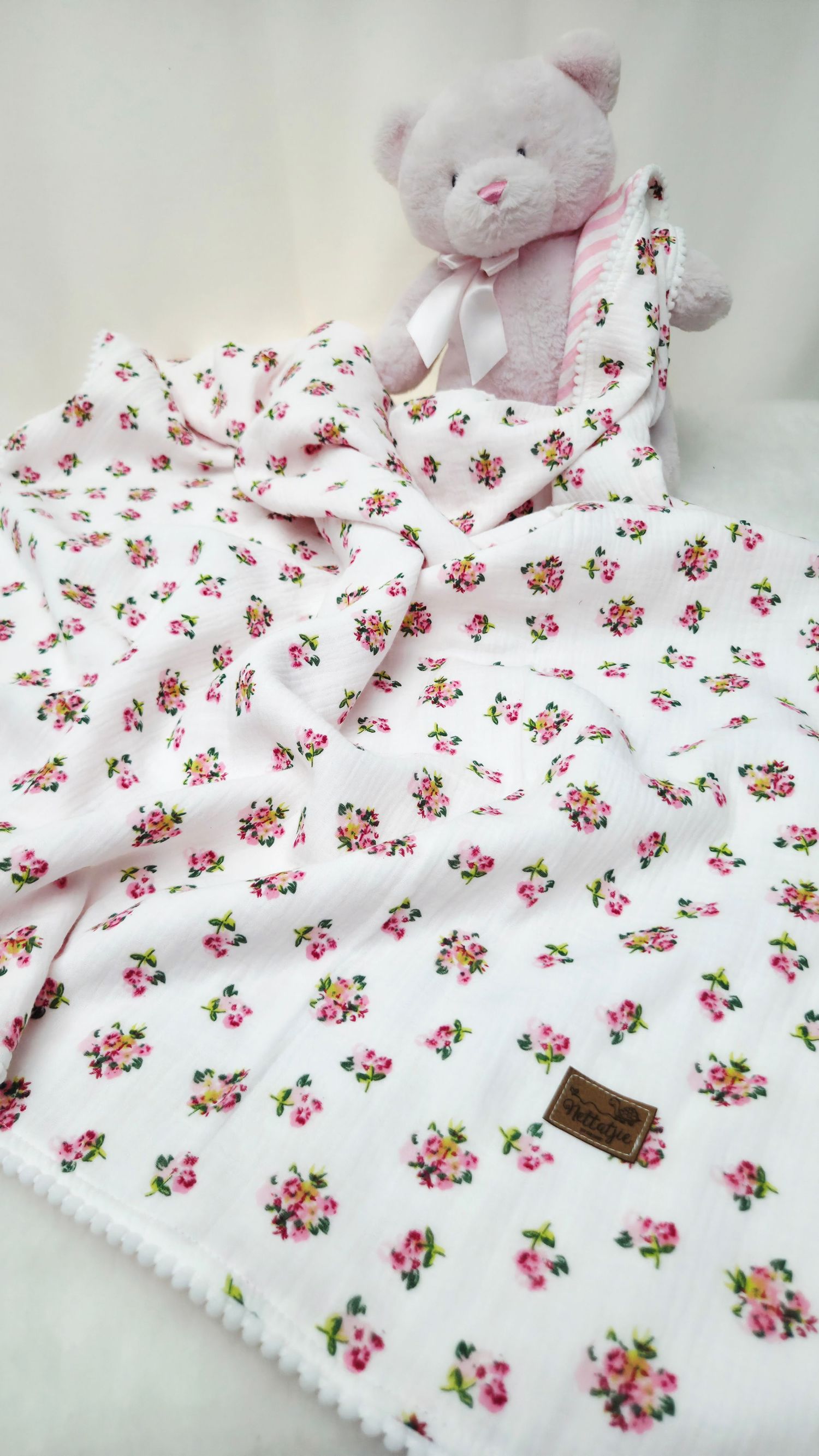 Pink floral cotton muslin baby blanket with pom-pom trim, draped beside a pink teddy bear with a satin ribbon.
