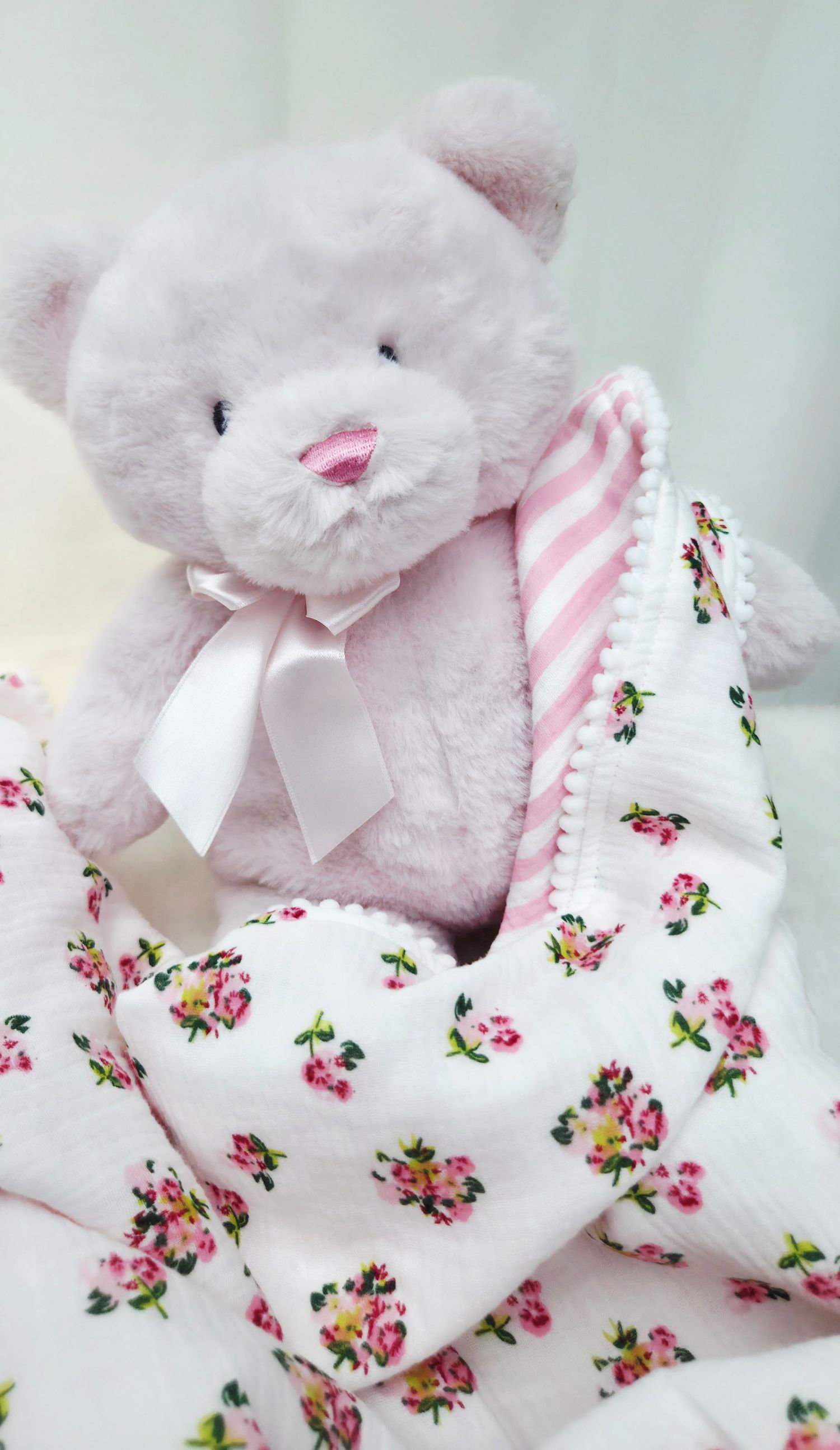 Soft pink plush teddy bear with a satin ribbon bow, wrapped in a floral baby blanket with pink striped lining and pom-pom trim.