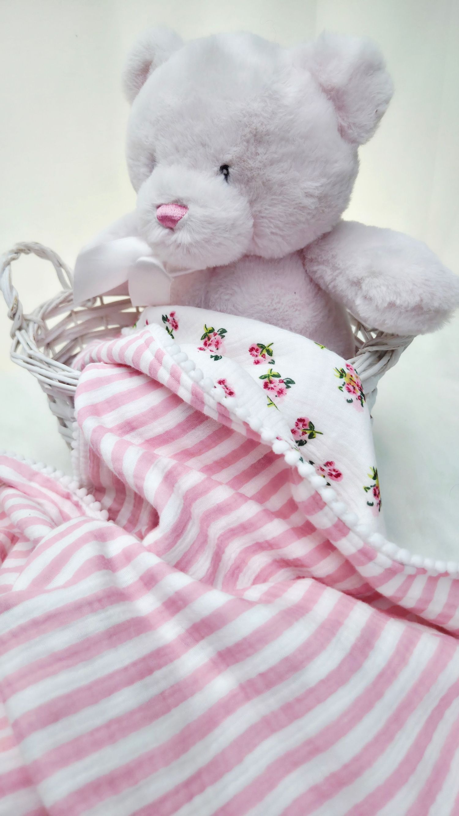 Pink plush teddy bear in a white wicker basket wrapped with a pink striped muslin baby blanket with floral lining and pom pom trim.