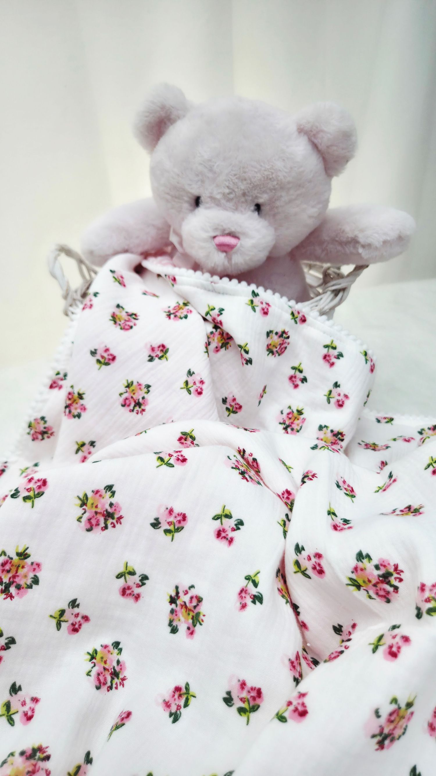 Soft pink teddy bear in a wicker basket draped with a white floral muslin baby blanket with pink rosebud print.